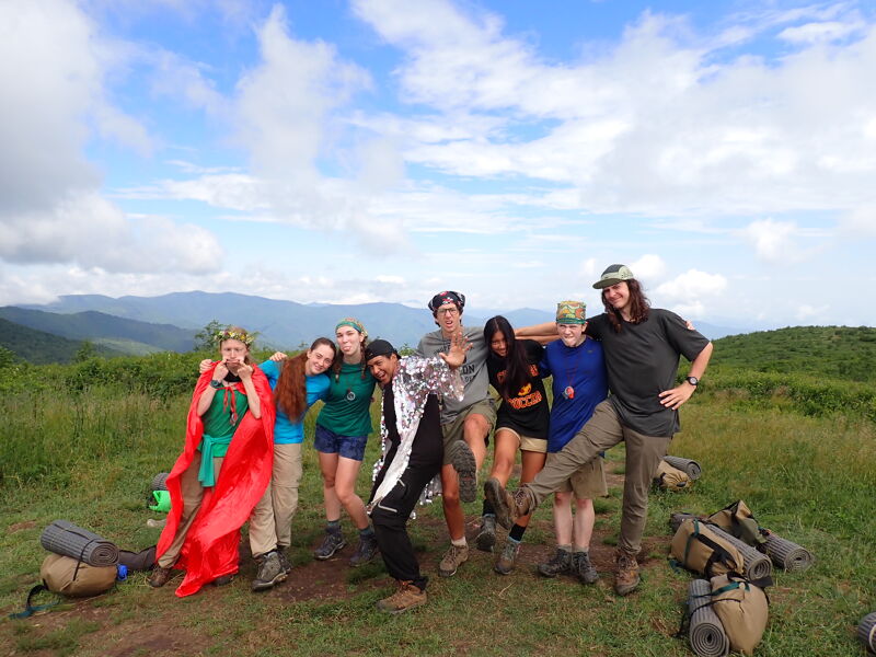 A group of nine young adults are standing together on a grassy hilltop, posing for a photo. They appear to be on a hiking trip, as some are wearing backpacks and hiking gear. The sky is partly cloudy, and there are mountains in the distance. The overall atmosphere is one of camaraderie and enjoyment of the outdoors.
